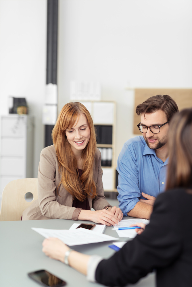 Young couple sitting in a meeting with an agent looking at a document together that she is presenting to them, view over the agents shoulder-1